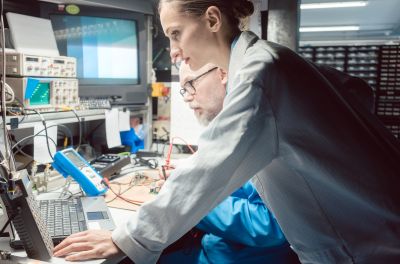 医療機器ODM/OEMサービス・モジュール開発のイメージ図。電子機器や測定器が並ぶ作業台で、2人の作業者が作業している様子。